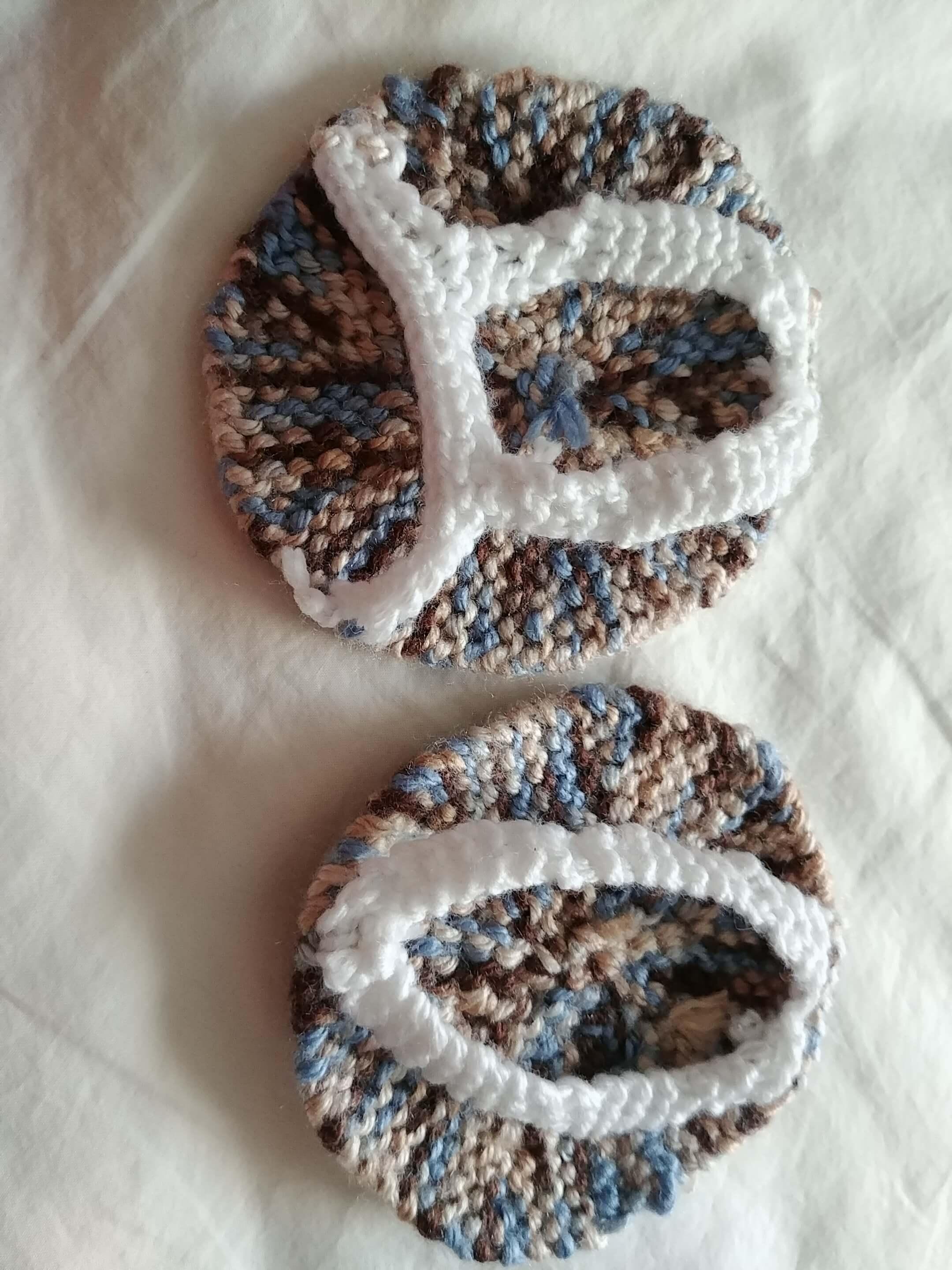 The headphone pads lying flat. The pads are made of blue and brown variegated yarn, with white knit straps. 